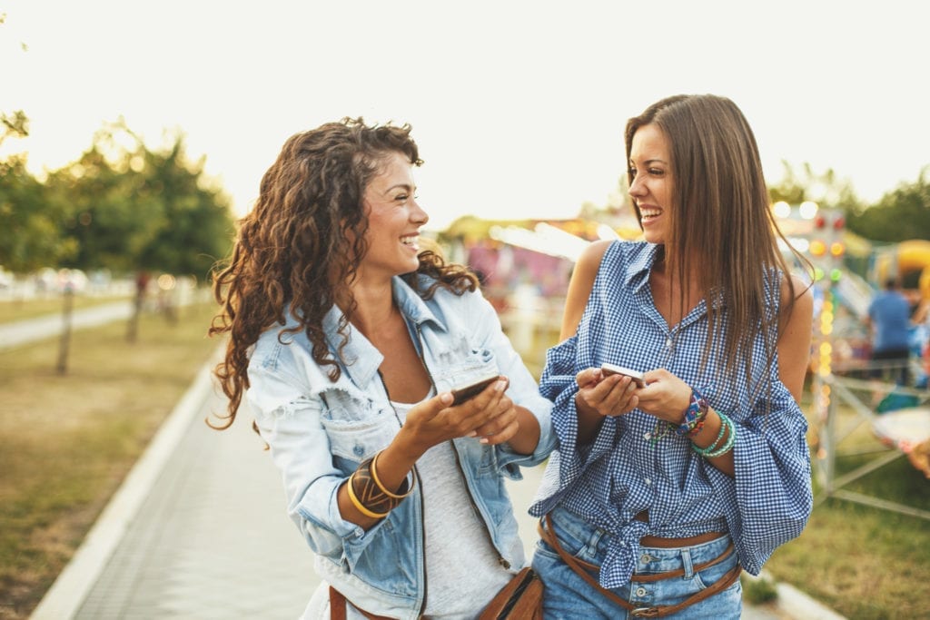   Two girls are outdoors, holding their smartphones and laughing.