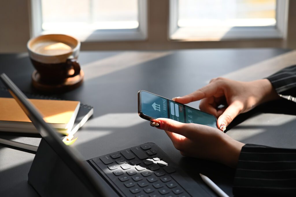 Cropped shot of executive woman holding smartphone in hands while doing online financial transaction by her smartphone in front computer tablet at the modern working desk.